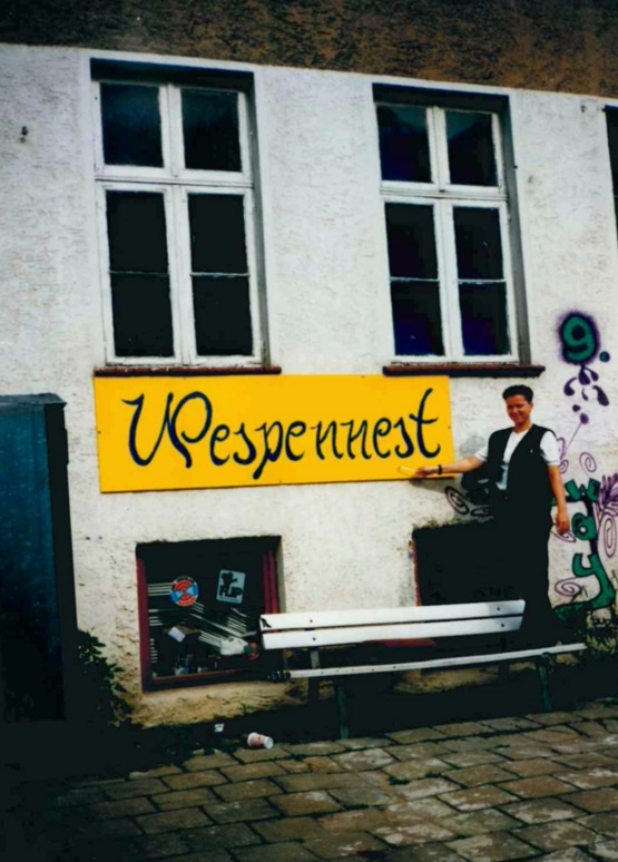 Außenansicht eines Hauses, eine Frau steht auf einer Gartenbank und zeit auf das Schild "Wespennest"